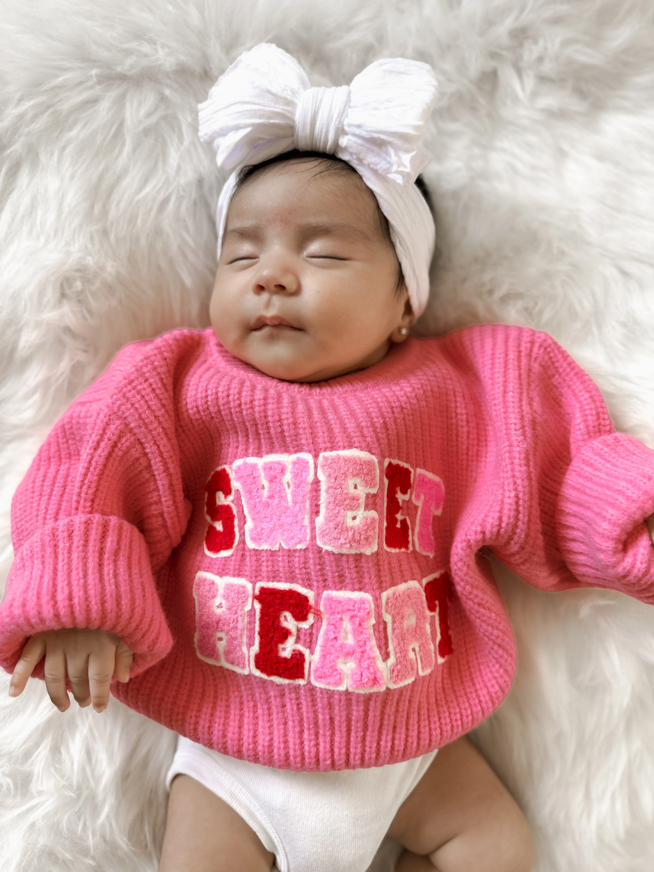 A serene baby girl in a pink "Sweet Heart" sweater and a white headband, resting on a fluffy white blanket.