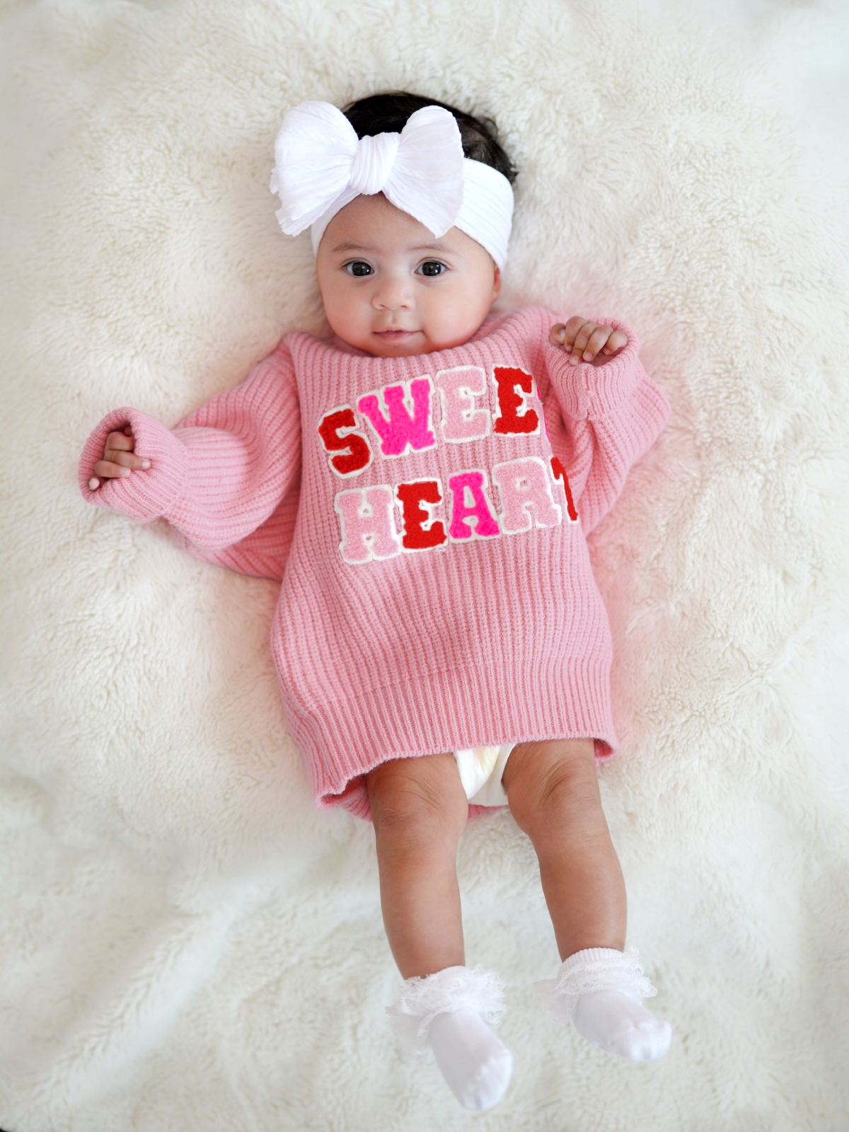 Baby girl in a pink sweater reading "SWEET HEART," wearing a white headband, lying on a fluffy blanket.