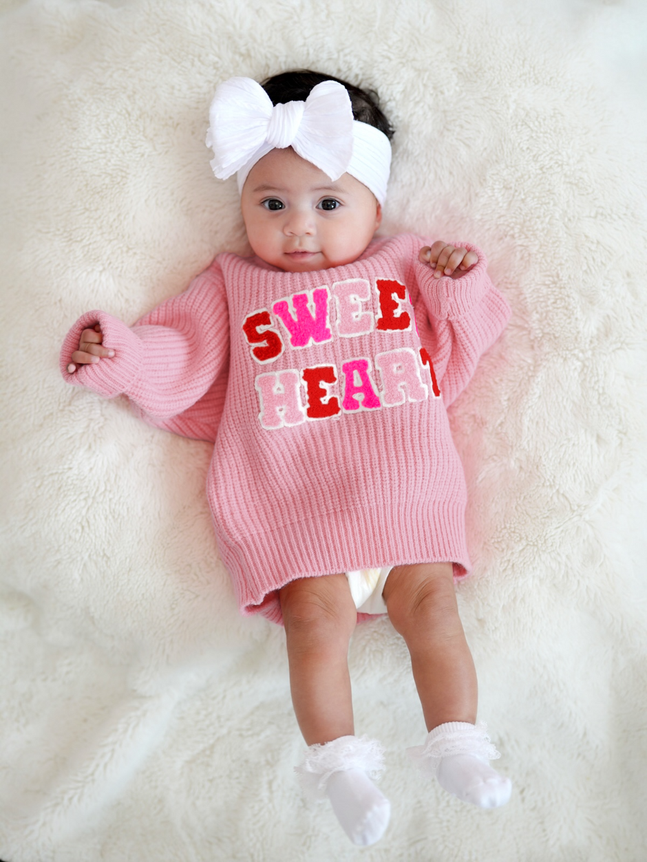 Baby girl in a pink sweater reading "SWEET HEART," wearing a white headband, lying on a fluffy blanket.