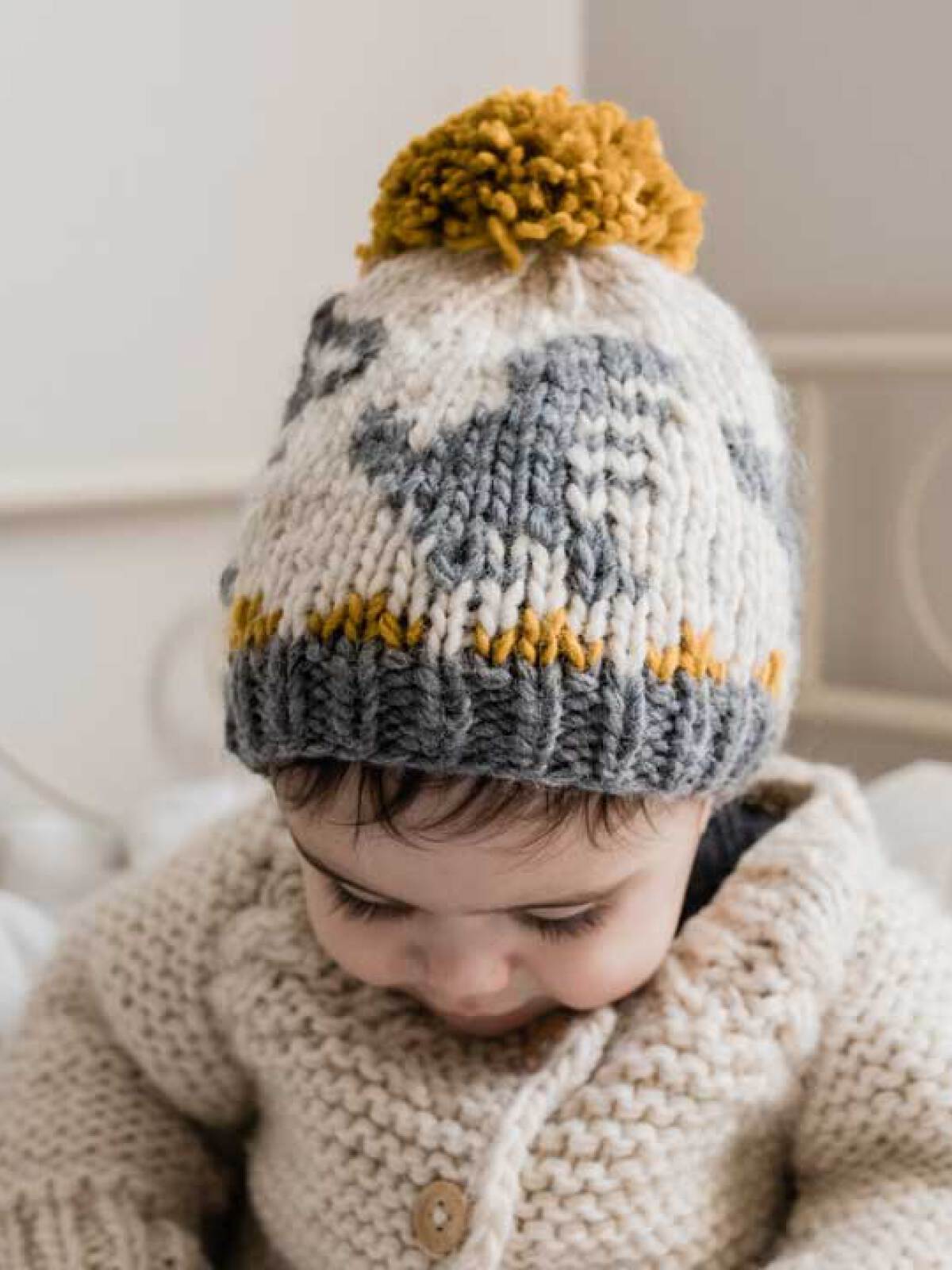 Baby wearing a cozy knit hat with a pom-pom, looking down while seated in a light-colored environment.