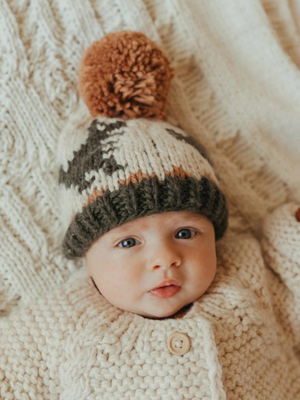 Baby in a knitted hat with a pom-pom, wearing a cozy sweater, resting on a textured blanket.