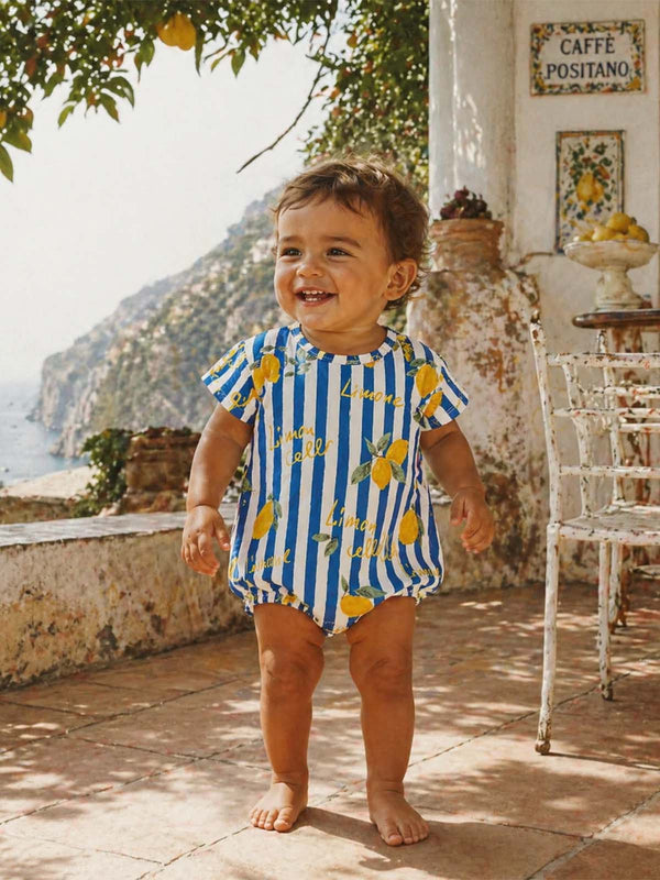 Smiling toddler in a lemon-patterned outfit stands outside in a sunlit, Mediterranean setting.