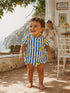 Smiling toddler in a lemon-patterned outfit stands outside in a sunlit, Mediterranean setting.
