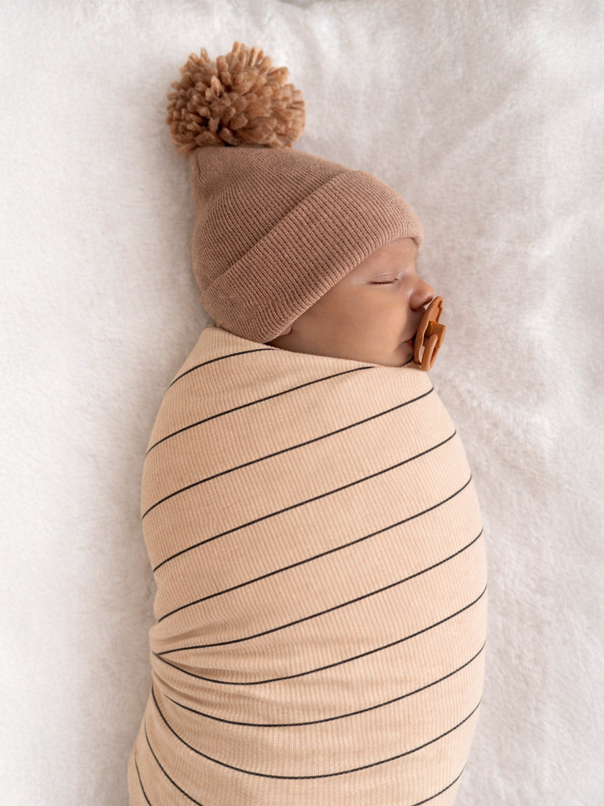 Sleeping baby swaddled in a striped blanket, wearing a cozy beige beanie with a pom-pom.