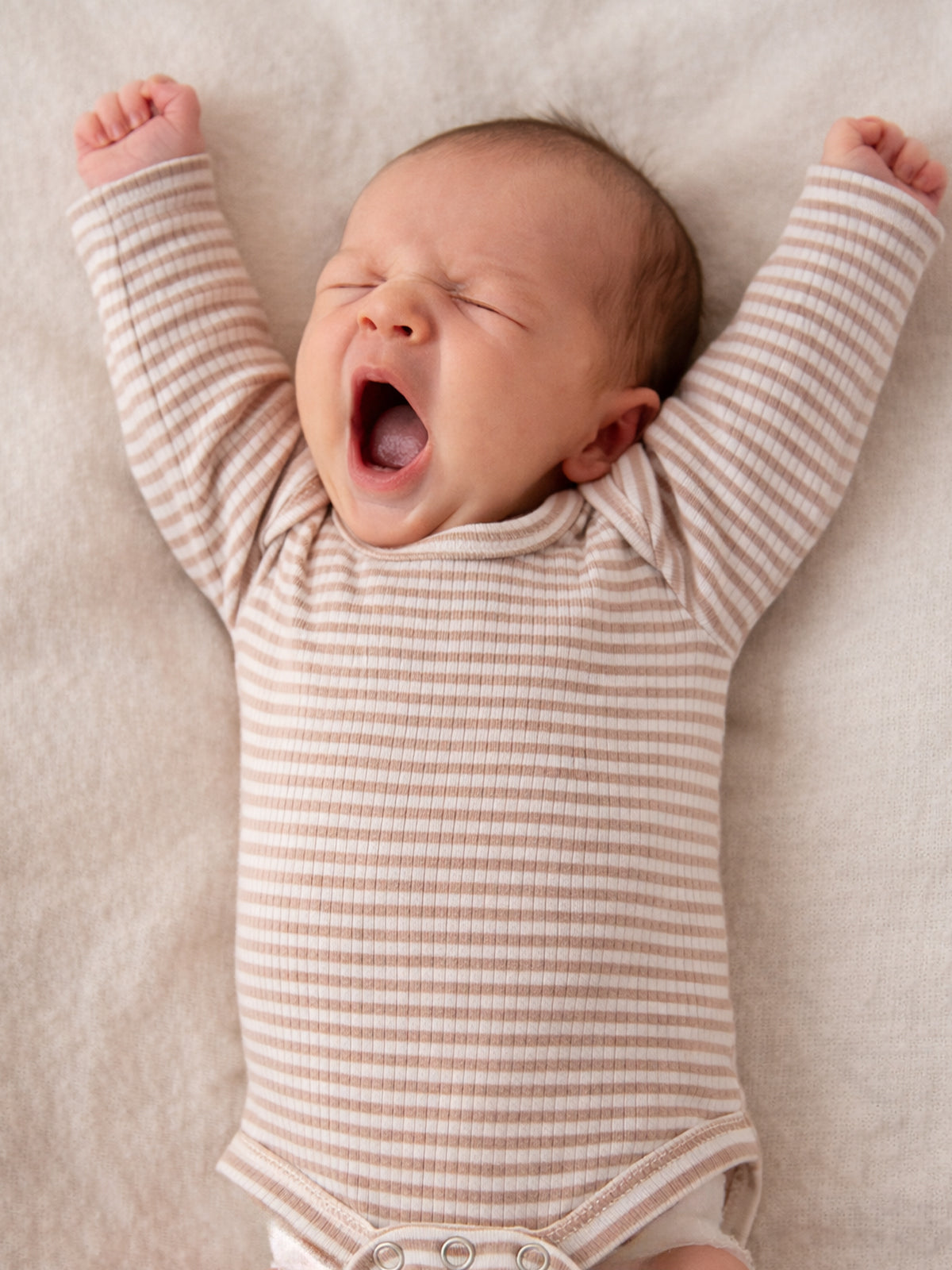 Infant in striped onesie yawning with arms raised against a soft, neutral background.
