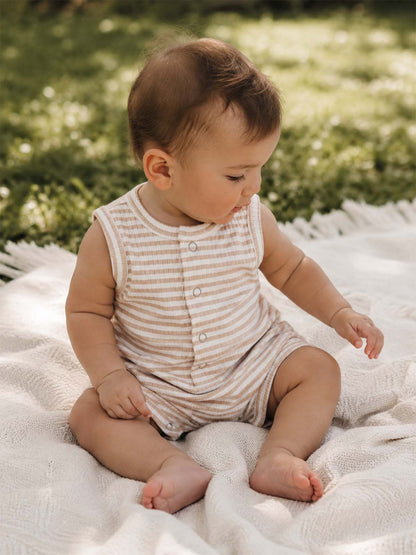 Baby wearing a striped onesie sits on a textured blanket in a grassy outdoor setting, absorbed in play.