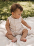 Baby wearing a striped onesie sits on a textured blanket in a grassy outdoor setting, absorbed in play.