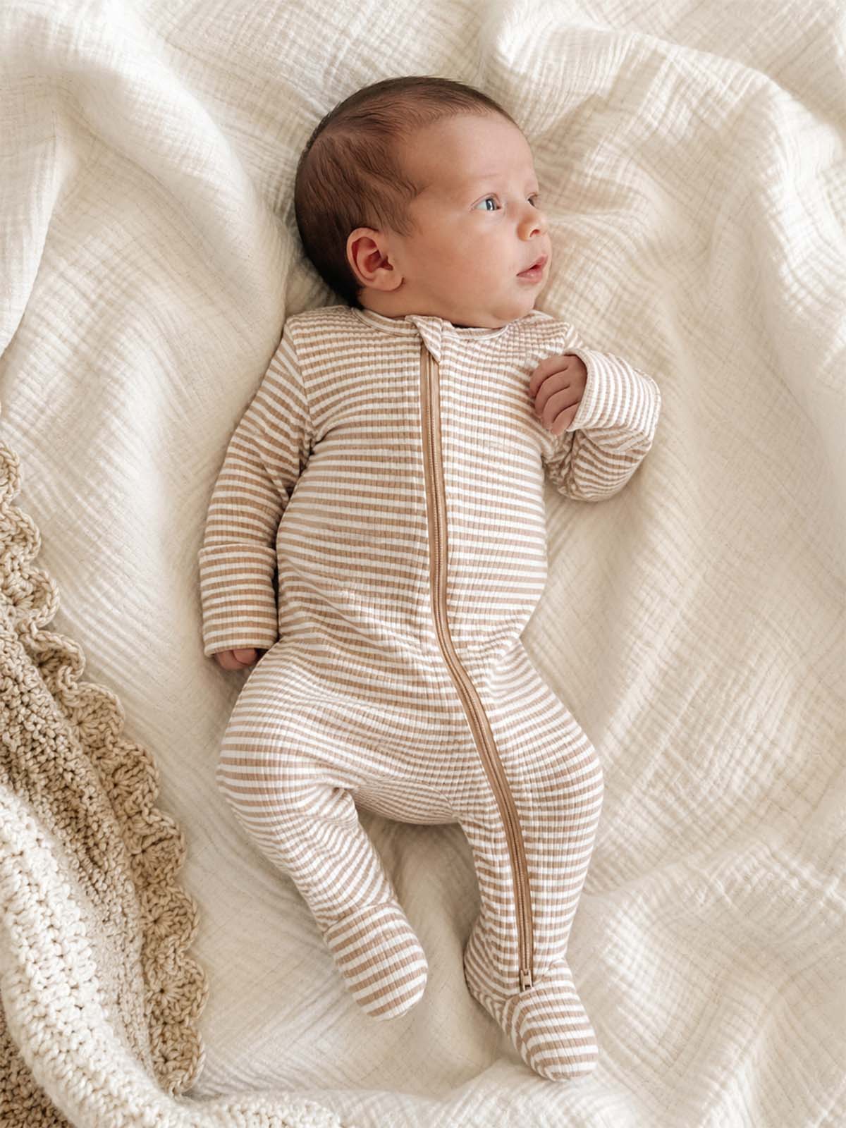 Baby in a striped sleeper lies on a soft blanket, looking up with a curious expression.
