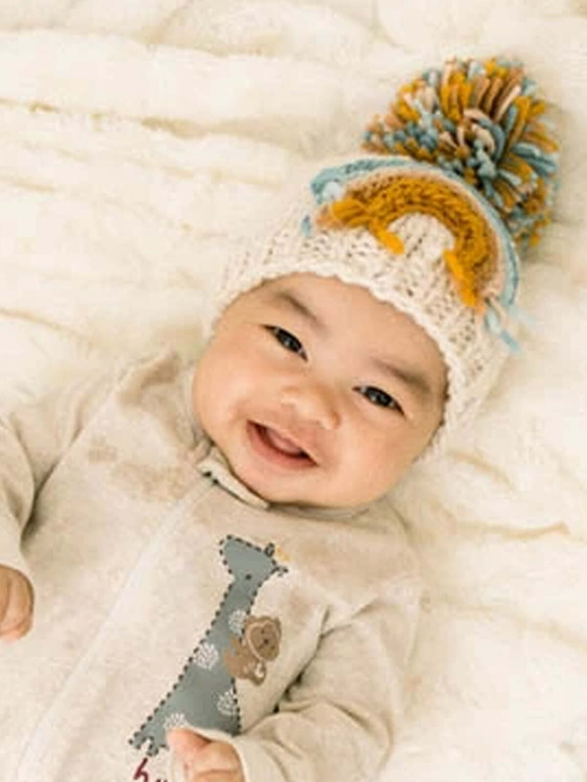 Happy baby wearing a knitted hat with pom-poms, laying on a soft blanket.