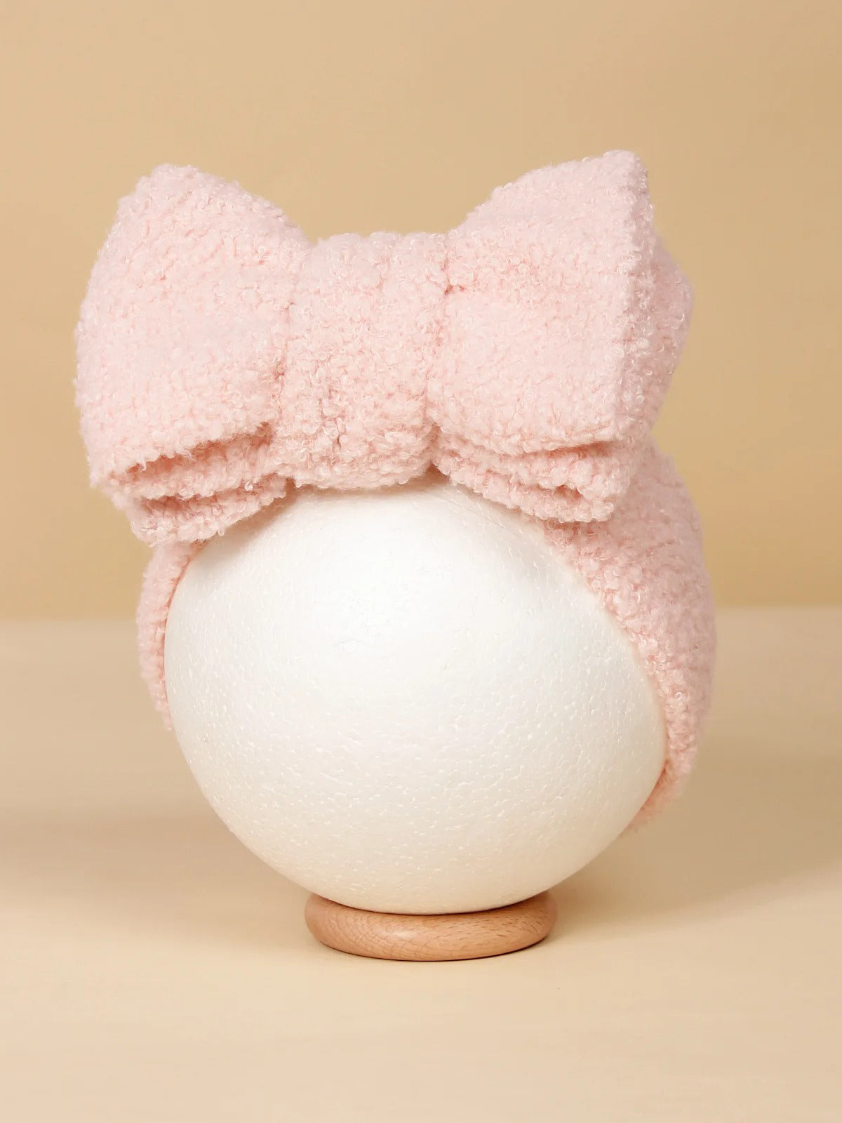 Pink fuzzy headband with a large bow displayed on a white sphere against a beige background.
