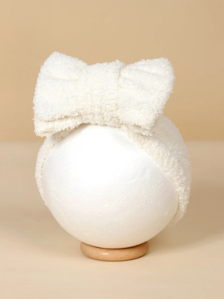 White fluffy headband with a large bow displayed on a round stand against a neutral background.