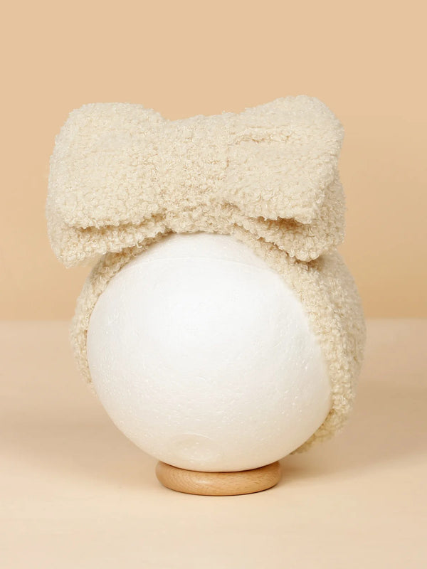 Cream-colored fuzzy headband with a large bow, displayed on a round pedestal against a beige background.