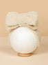 Cream-colored fuzzy headband with a large bow, displayed on a round pedestal against a beige background.