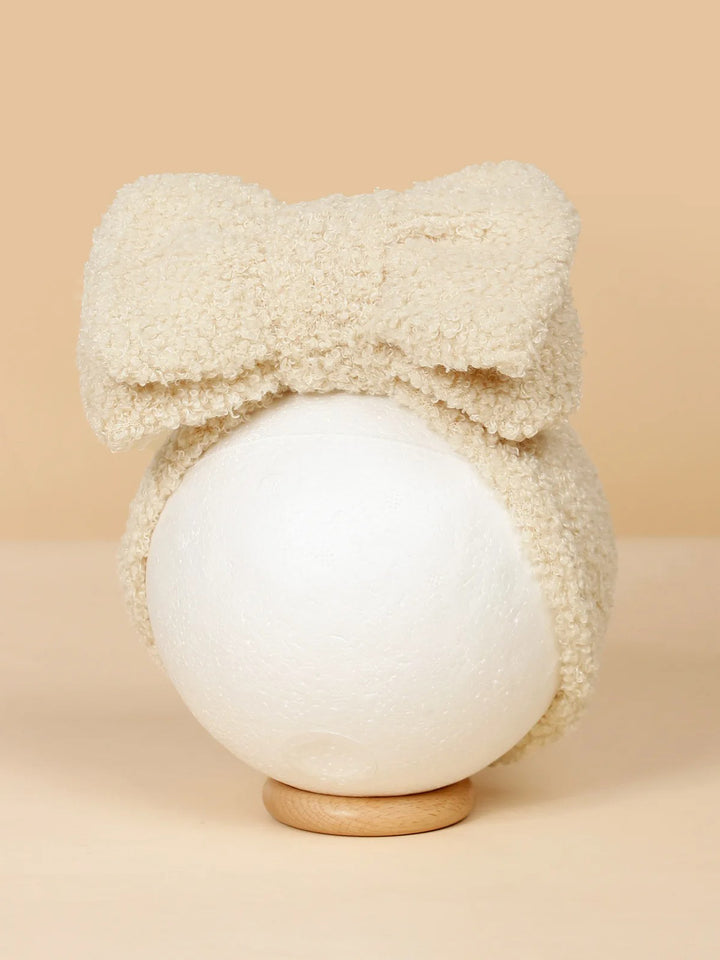 Cream-colored fuzzy headband with a large bow, displayed on a round pedestal against a beige background.
