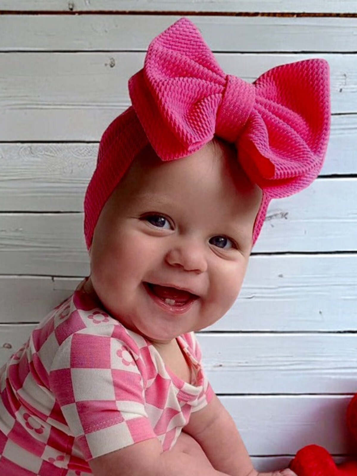 Smiling baby girl wearing a pink checkered outfit and a large pink bow, sitting on a wooden surface.