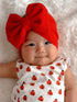 Smiling baby girl in a white ladybug outfit and a large red bow on a soft, textured background.