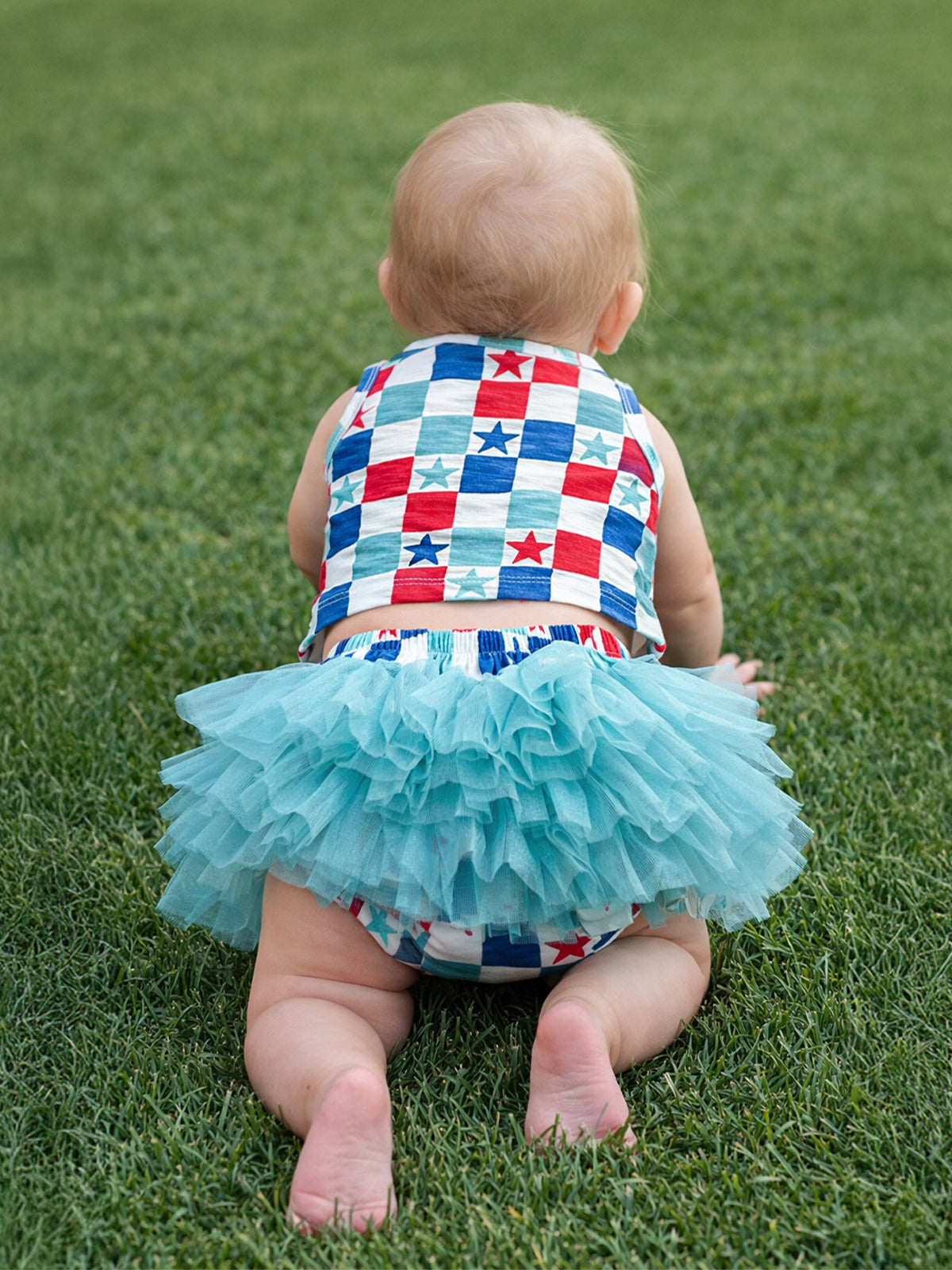 Baby crawling on grass, wearing a colorful star-patterned outfit and a fluffy turquoise tutu.