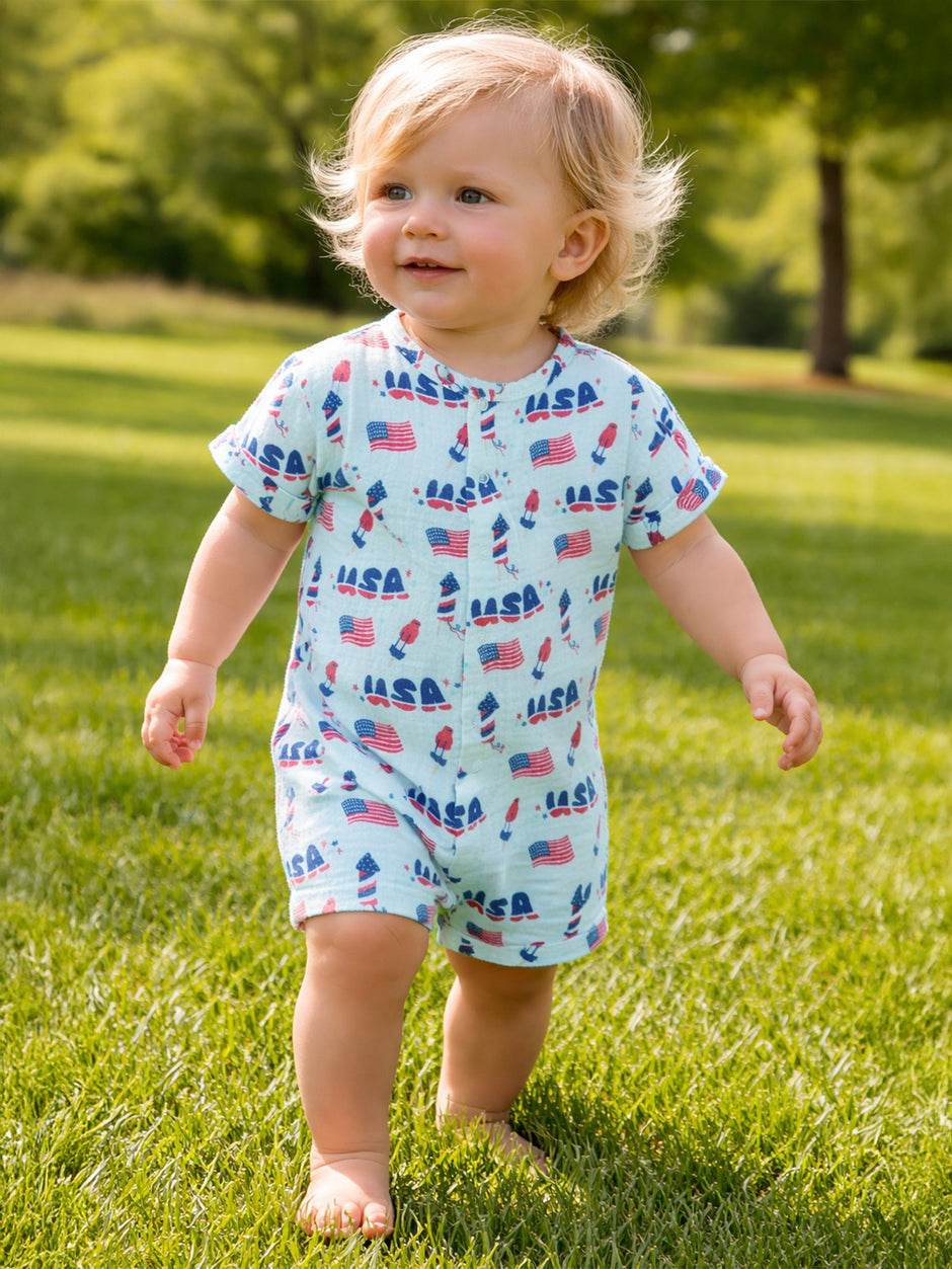 Toddler with curly blonde hair wearing a patterned romper, walking on green grass outdoors.