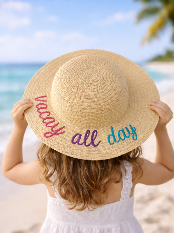 Child wearing a straw sun hat with "Vacation All Day" text, standing on a beach with ocean in the background.