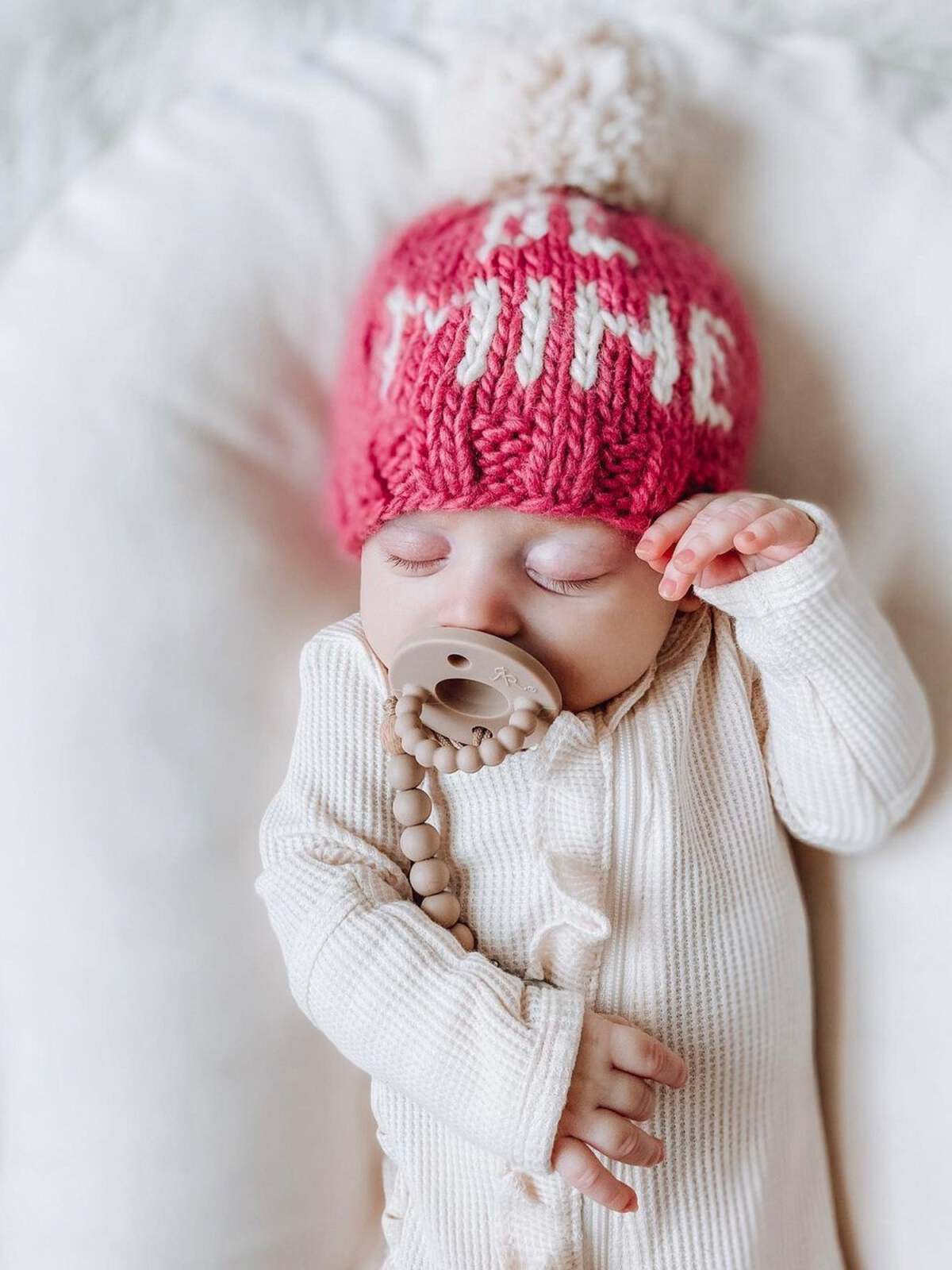 Baby peacefully sleeping in a pink knitted hat with pom pom, wearing a cream-colored outfit and soothing pacifier.