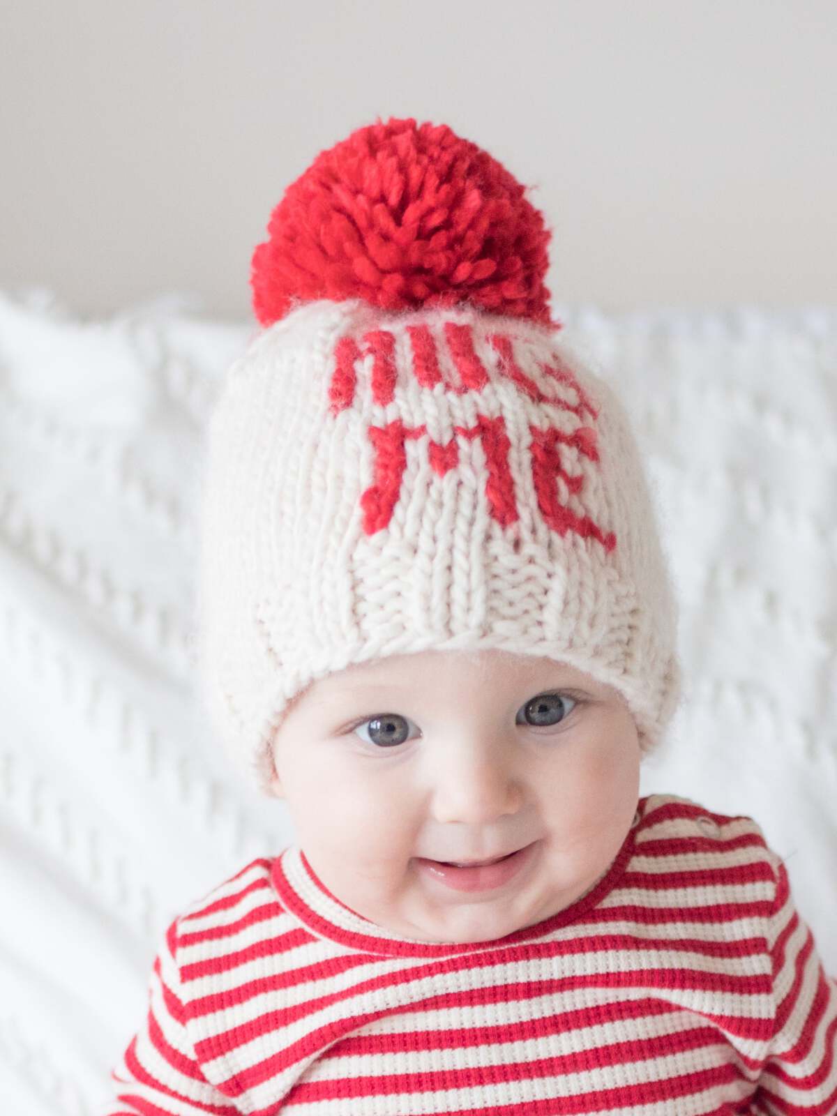 Smiling baby wearing a knitted hat with a red pom-pom and "HUG ME" text, dressed in a striped red and white outfit.