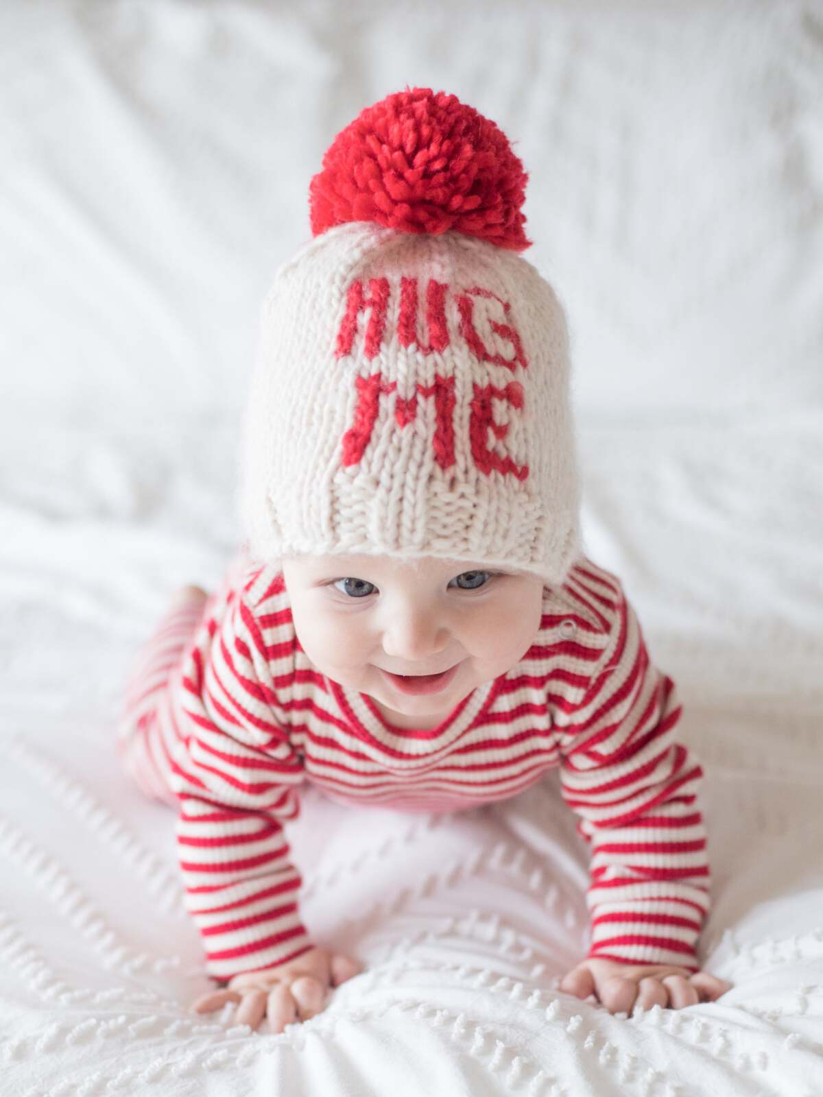 Baby in a knitted "Hug Me" hat crawling on a white blanket, wearing a red and white striped outfit.