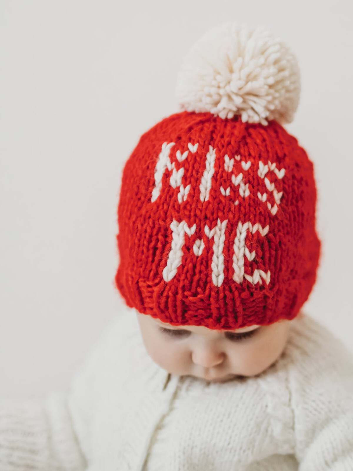 Baby wearing a red knitted hat with "KISS ME" in white letters and a fluffy pom-pom on top.