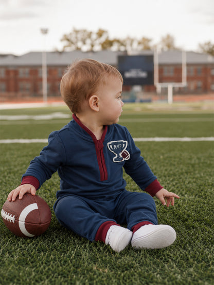 Toddler in navy MVP outfit sitting on grass, holding a football with a blurred football field in the background.