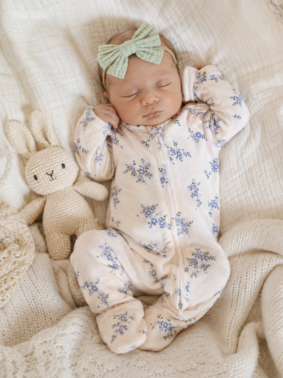 Sleeping baby in floral onesie with green bow, cuddling a knitted bunny toy on a cozy blanket.
