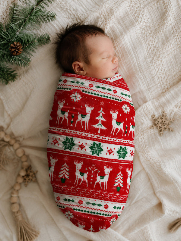 Newborn swaddled in festive red fabric with deer and trees, resting on a cozy blanket with pine branches nearby.