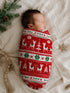 Newborn swaddled in festive red fabric with deer and trees, resting on a cozy blanket with pine branches nearby.