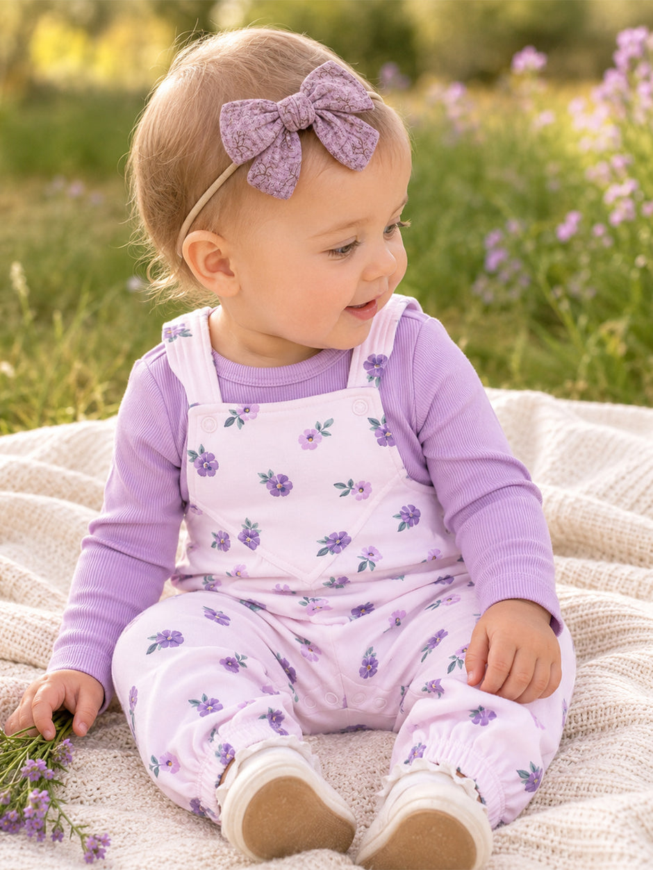 Toddler girl with a lavender bow, wearing floral overalls, sitting on a blanket outdoors with flowers around her.
