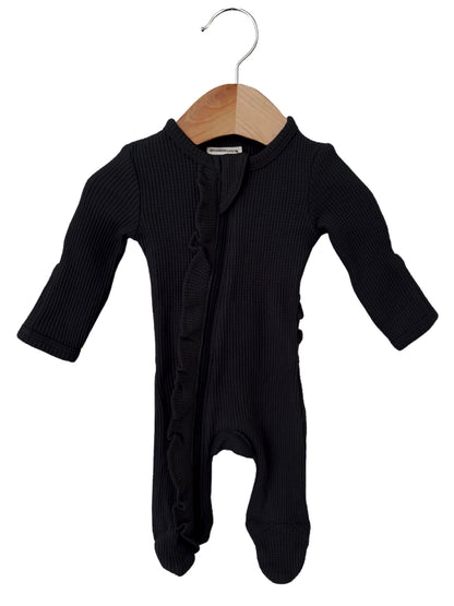 Black baby onesie with long sleeves and ruffle details, hanging on a wooden hanger against a white background.