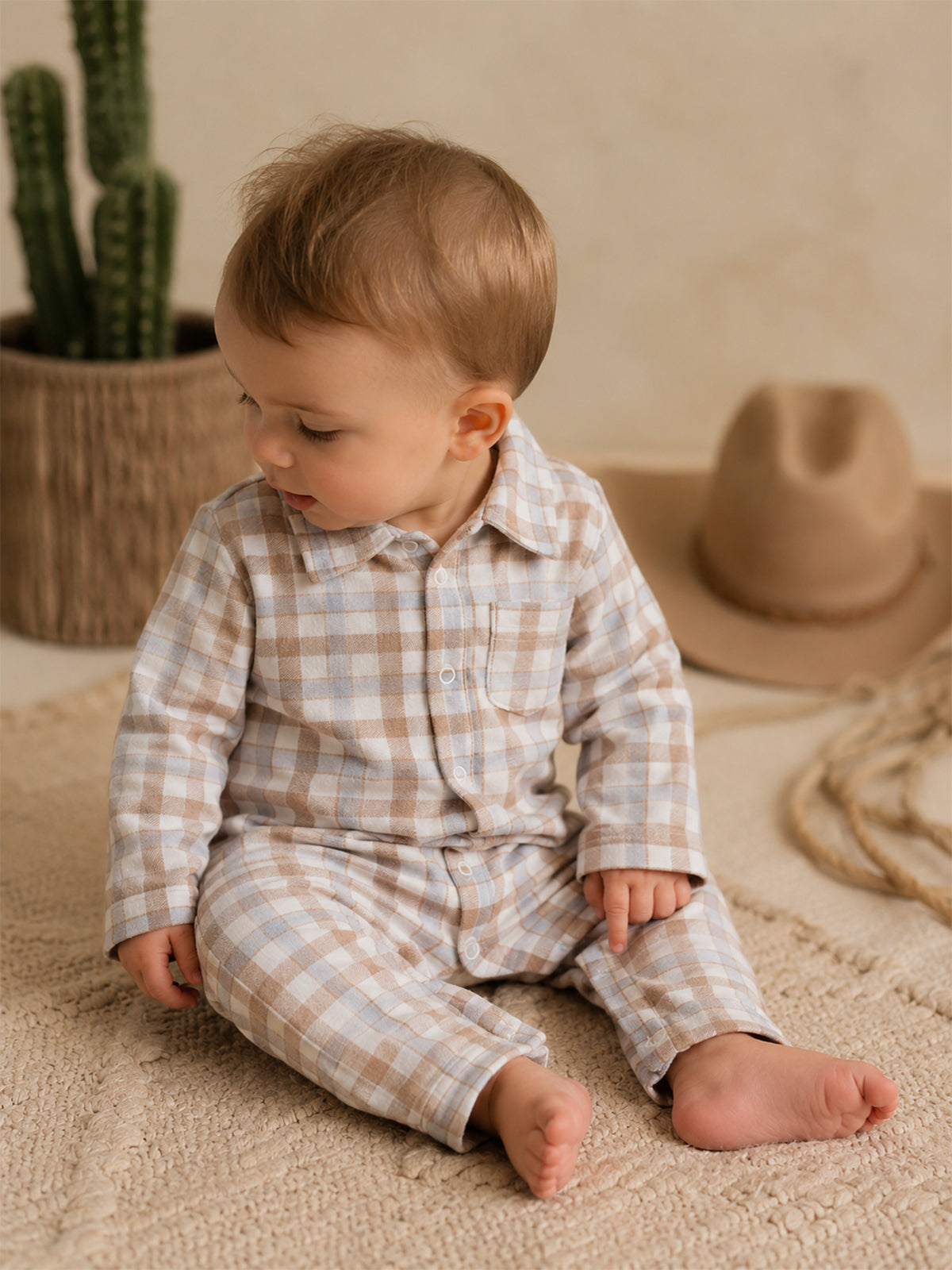 Toddler in a plaid outfit sits on a textured mat, with a cactus and hat in the background.