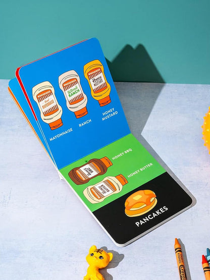 Colorful children's book page showing condiments and pancakes with a toy cat and crayons nearby.