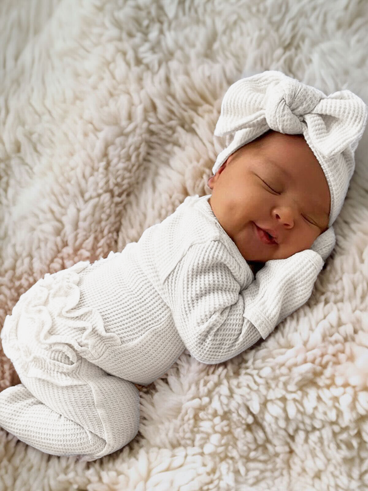 Smiling baby in a white outfit with a bow, peacefully resting on a soft, fluffy blanket.