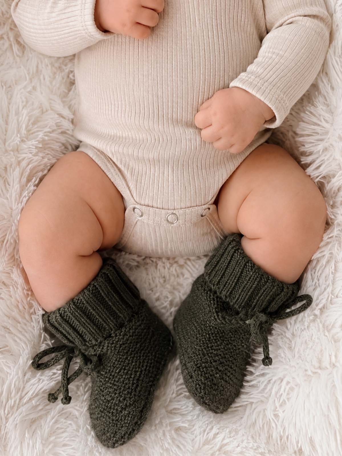 Baby in a ribbed onesie with chunky green knitted booties, resting on a fluffy white blanket.