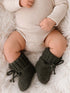 Baby in a ribbed onesie with chunky green knitted booties, resting on a fluffy white blanket.