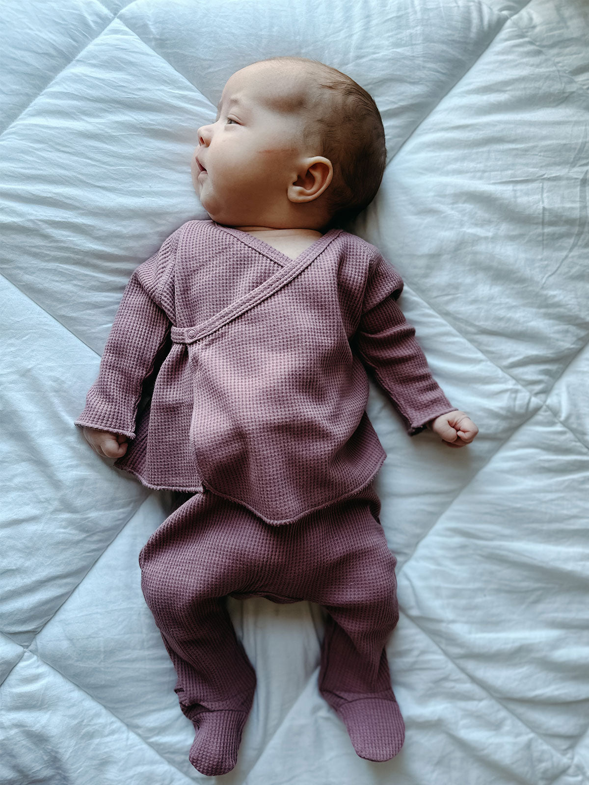 Baby in a lavender outfit resting on a white quilt, looking to the side with short hair.
