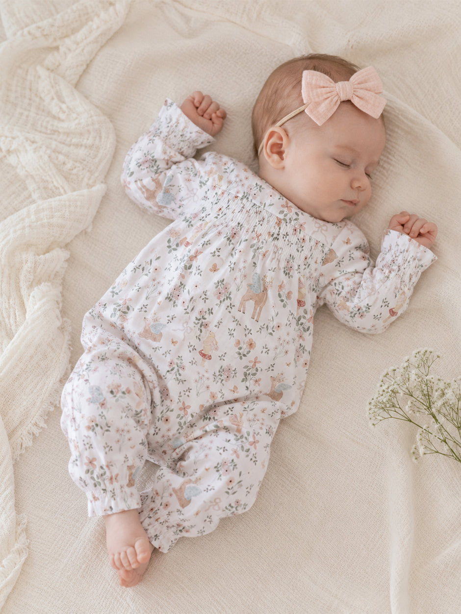 Sleeping baby in a floral onesie with a pink bow, resting on a soft blanket with delicate flowers nearby.