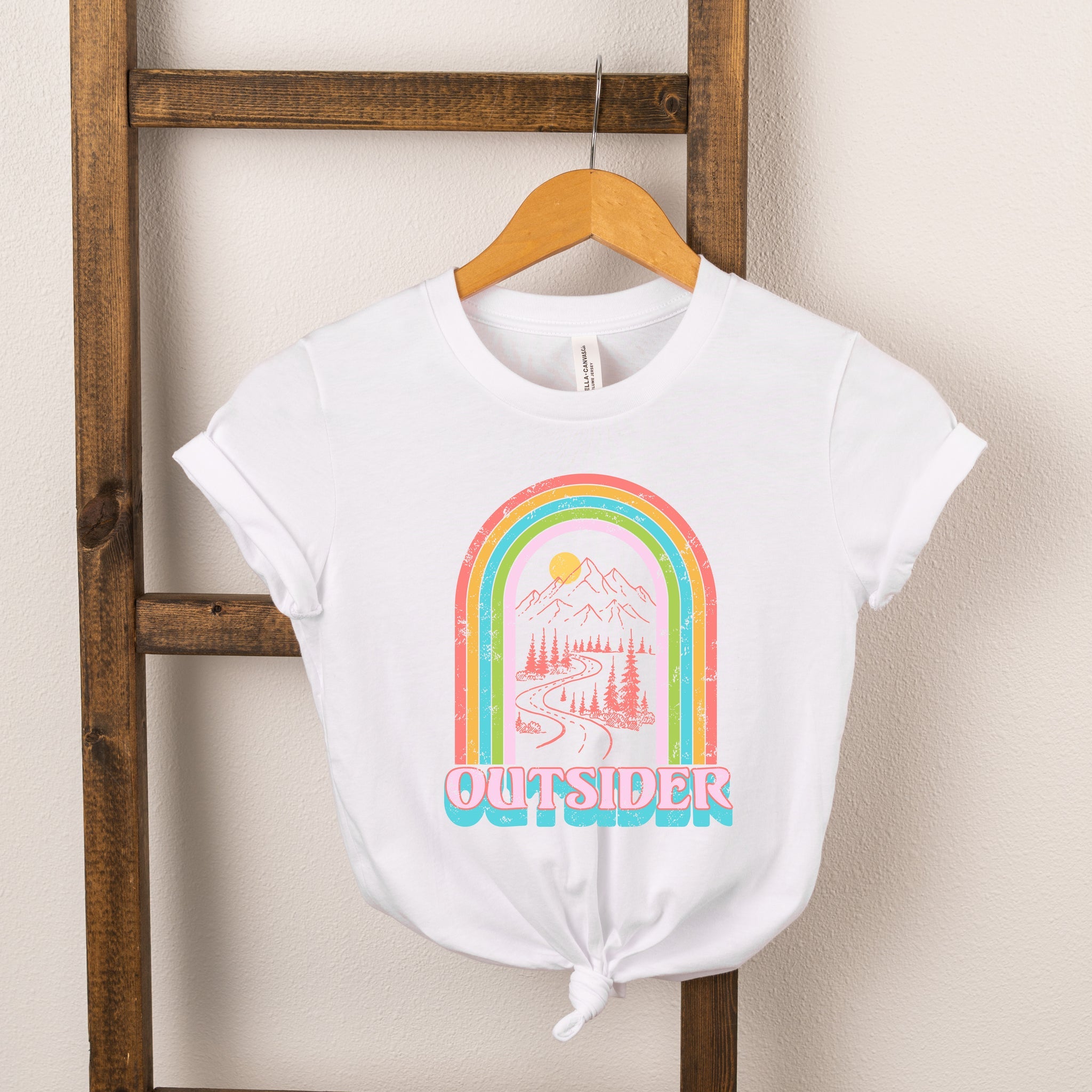 White t-shirt featuring a colorful rainbow design and "OUTSIDER" text, hung on a wooden ladder.