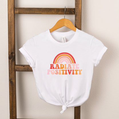 White t-shirt with "Radiate Positivity" and a rainbow graphic, hanging on a wooden ladder.