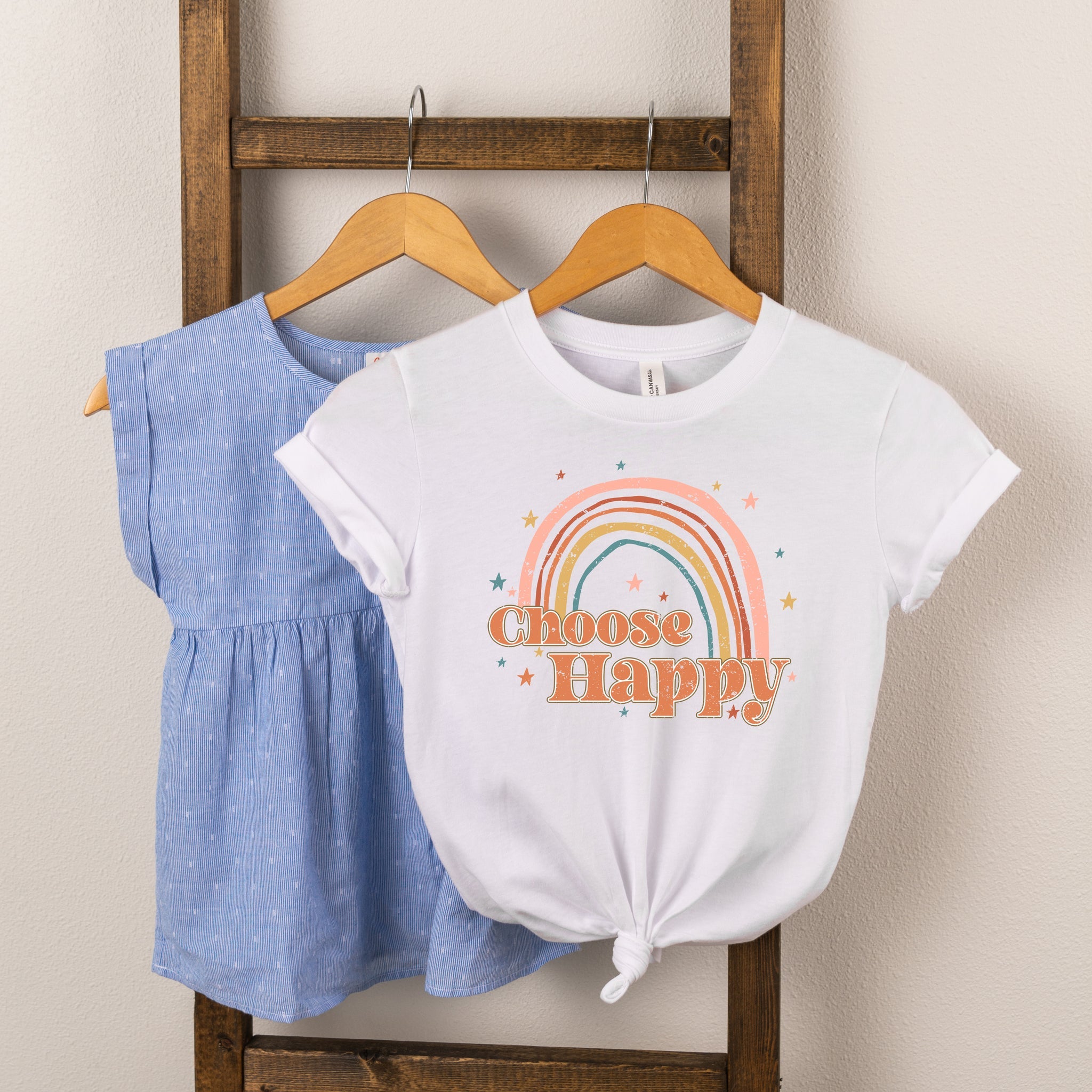 Two children's shirts displayed on a wooden hanger: a blue top and a white shirt with a rainbow and "Choose Happy" print.