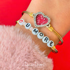 Two bracelets on a wrist: one with a sparkling heart charm, the other spells "I LOVE U MORE" in beads.