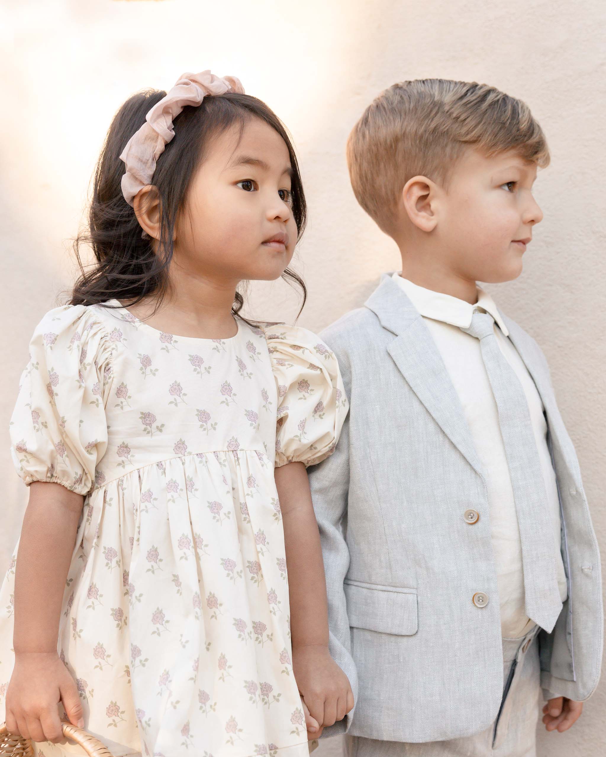 Two children, a girl in a floral dress and a boy in a suit, standing side by side against a neutral background.