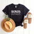 Black t-shirt with "BONUS mama" text, a tan hat, and sandals on a white wooden background.