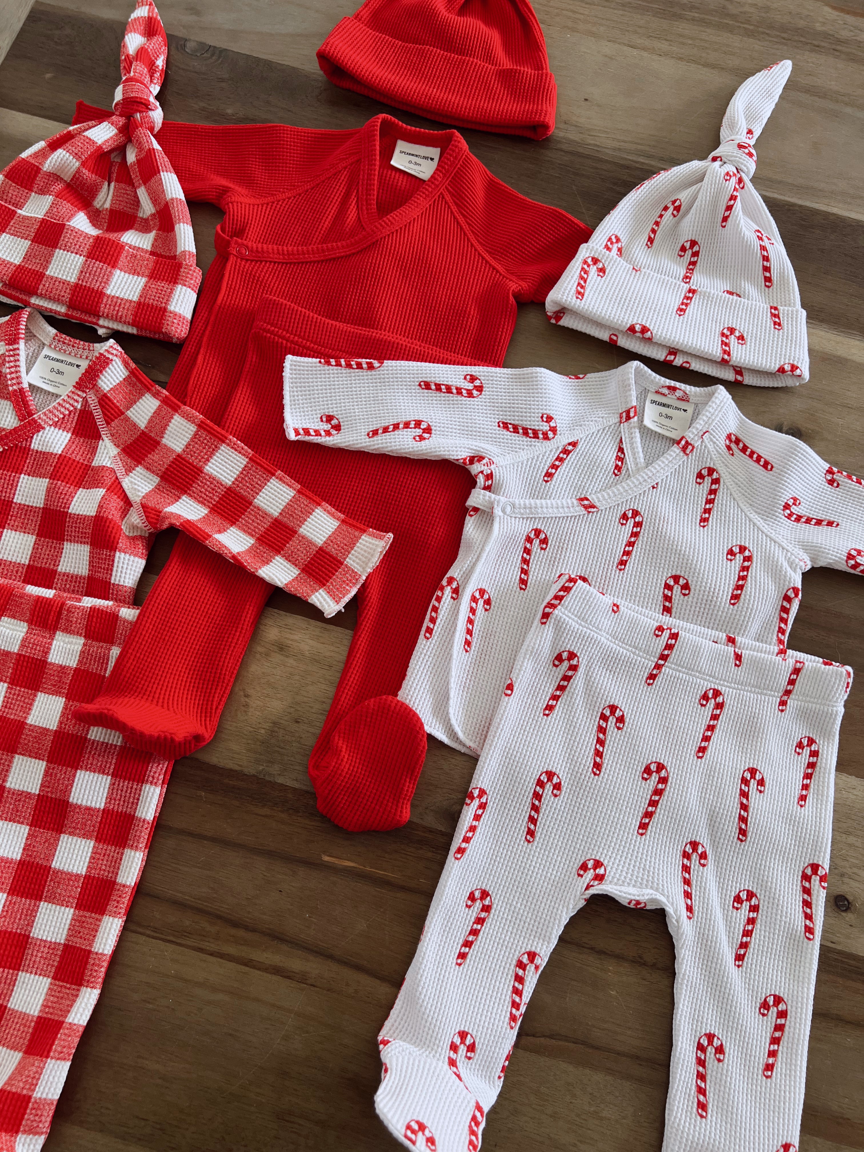 Colorful holiday baby clothes including red and candy cane patterned outfits and hats on a wooden surface.