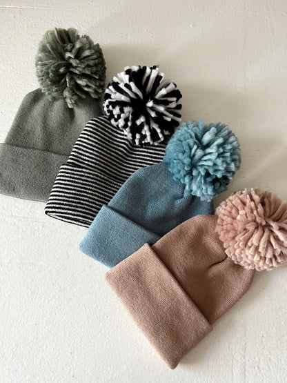 Four beanies with pom-poms displayed on a light surface: gray, striped black and white, blue, and pink.