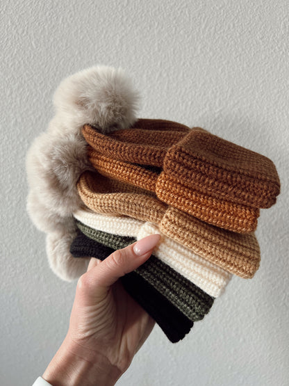 Hand holding a stack of colorful crocheted hats with pom-poms against a textured white background.
