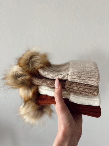 A hand holding a stack of knit hats in beige, brown, cream, and rust colors, featuring fluffy pom-poms.